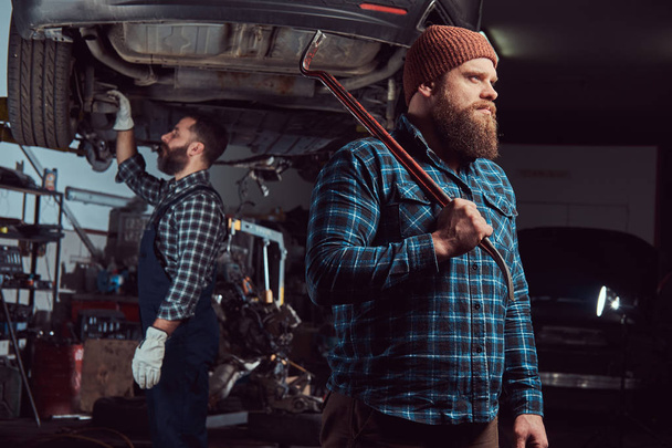 Дві бородаті жорстокі механіки ремонтують машину на ліфті в гаражі
.  - Фото, зображення