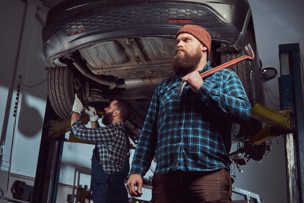 Twee bebaarde brute mechanica repareren een auto op een lift in de garage.  - Foto, afbeelding