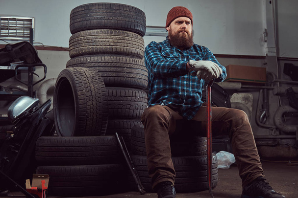 ein bärtiger Mechaniker in Uniform sitzt auf alten Autoreifen in der Garage. - Foto, Bild
