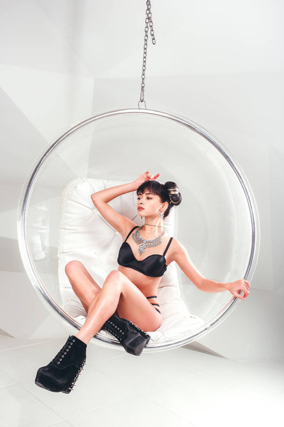 Beautiful slim model woman in underwear posing siting in suspended transparent chair in white studio. - Photo, Image