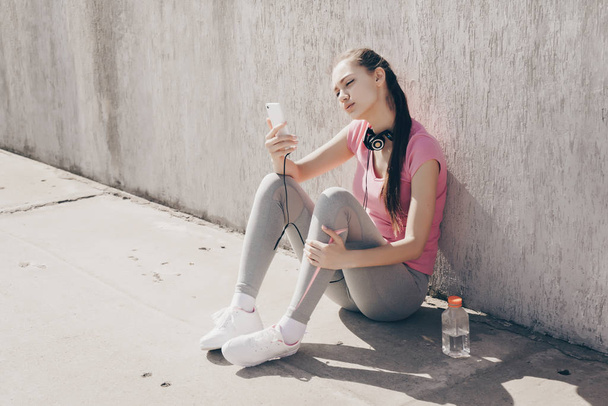 young athletic girl sitting on the ground outdoors, resting after workout and looking into smartphone - Valokuva, kuva