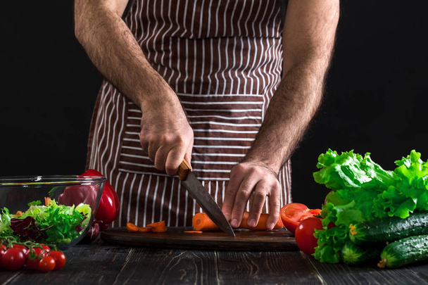 Young home cook man in apron slicing carrot with kitchen knife. Mens hands cut the carrot to make a salad on black background. Healthy food concept - Foto, immagini