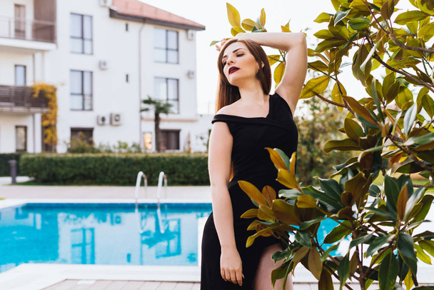 a luxurious rich woman in a black dress walking through her garden by the blue pool - Foto, afbeelding