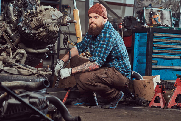 A brutal tattooed bearded mechanic specialist repairs the car engine which is raised on the hydraulic lift in the garage. Service station. - Photo, Image