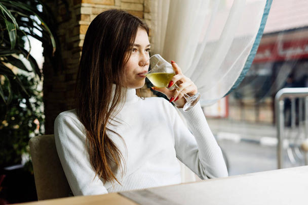 tmavovlasá hezká dívka v bílém svetru sedí v kavárně a pije bílé víno - Fotografie, Obrázek