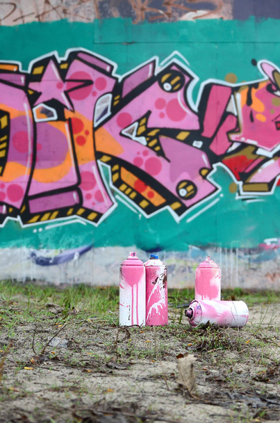 A few used paint cans lie on the ground near the wall with a beautiful graffiti painting in pink and green colors. Street art concept - Photo, Image
