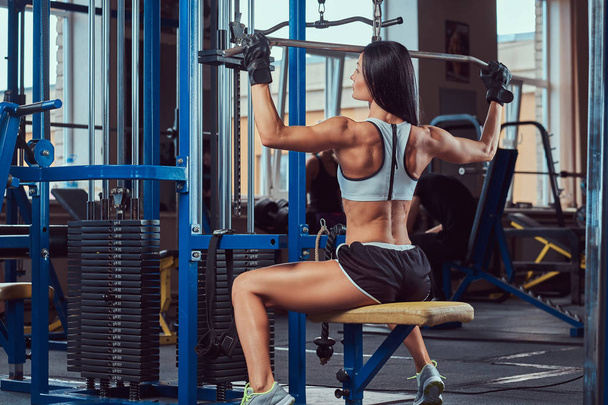 Красивая сексуальная атлетичная юная брюнетка в спортивной форме тренируется, накачивая мышцы задних латов в спортзале
. - Фото, изображение
