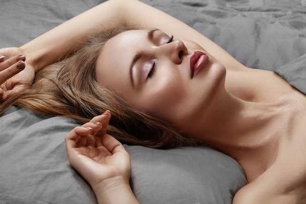 Bella mujer durmiendo mientras está acostada en la cama con comodidad. Dulces sueños. Modelo sexy con cabello largo rizado relajante en sábanas grises
. - Foto, Imagen