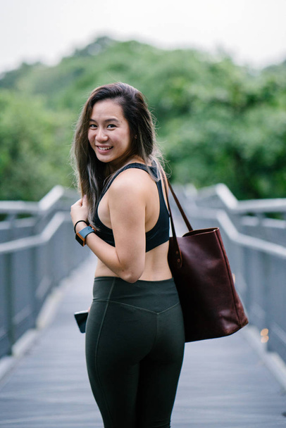 Portrait of an attractive, young Chinese Asian woman dressed in sporty, active wear with bag in green public park in Singapore during the day on the weekend. - 写真・画像