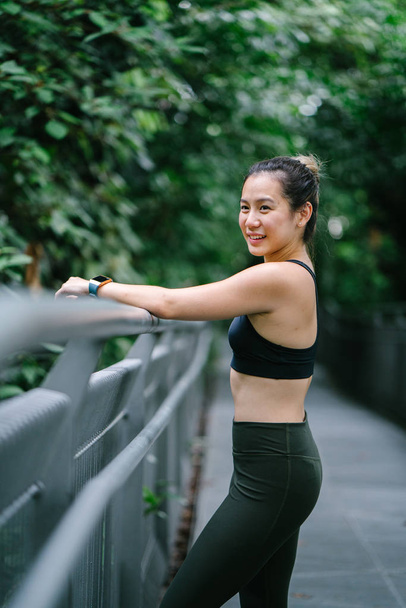 Portrait of an attractive, young Chinese Asian woman dressed in sporty, active wear on a bridge in a lush, green public park in Singapore during the day on the weekend. - Foto, imagen