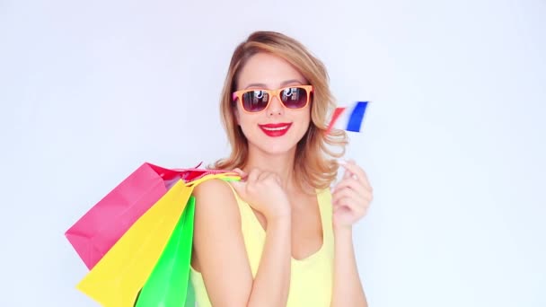 Chica pelirroja joven en gafas de sol con bolsas de compras y bandera francesa sobre fondo blanco
 - Metraje, vídeo