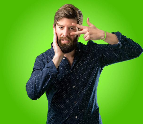 Jeune homme hipster avec une grosse barbe regardant la caméra à travers ses doigts dans le geste de victoire clin d'oeil et souffler un baiser sur fond vert
 - Photo, image