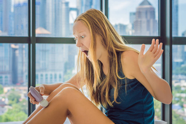 Young woman siting on bed at home and doing epilation with epilator on legs and felling pain. Window overlooking big city skyscrapers on background  - Photo, image