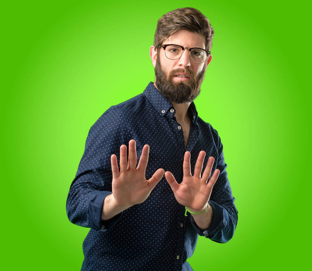 Jonge hipster man met grote baard geërgerd met slechte houding maken stopbord met hand, zegt Nee, uiting van veiligheid, defensie of beperking, misschien duwen over groene achtergrond - Foto, afbeelding