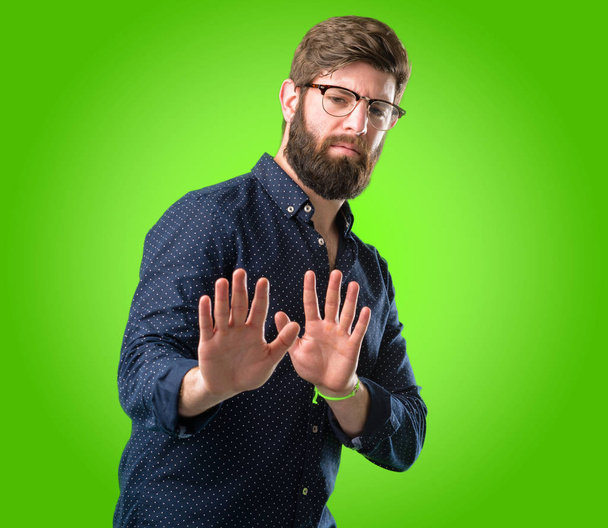 Молодой хипстер с большой бородой раздражен плохим отношением делает знак стоп с рукой, говоря нет, выражая безопасность, защиту или ограничение, может быть, толкая через зеленый фон
 - Фото, изображение