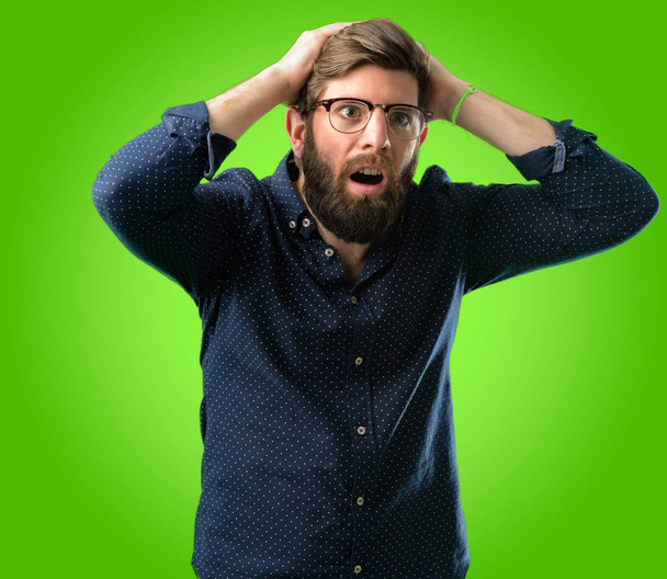 Молодой хипстер с большой бородой напряженно держа руки на голове, напуганный в панике, крича на зеленом фоне
 - Фото, изображение
