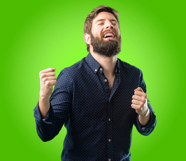 Молодой хипстер с большой бородой счастлив и взволнован празднованием победы, выражая большой успех, силу, энергию и положительные эмоции. Празднует новую работу радостно на зеленом фоне
 - Фото, изображение