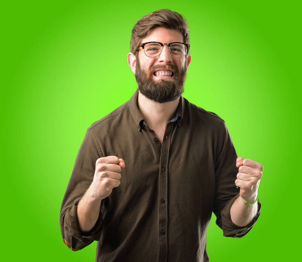 Молодой хипстер с большой бородой счастлив и взволнован празднованием победы, выражая большой успех, силу, энергию и положительные эмоции. Празднует новую работу радостно на зеленом фоне
 - Фото, изображение