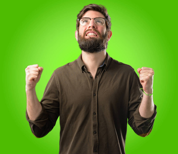 Joven hombre hipster con barba grande feliz y emocionado celebrando la victoria expresando gran éxito, poder, energía y emociones positivas. Celebra nuevo trabajo alegre sobre fondo verde
 - Foto, imagen