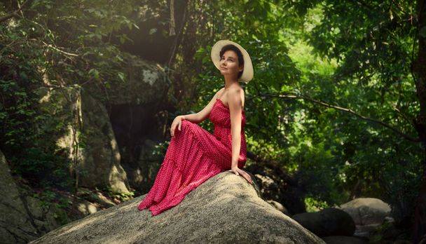 Ein Mädchen mit Hut sitzt auf einem Felsen im Dschungel. Mädchen im roten Kleid. - Foto, Bild