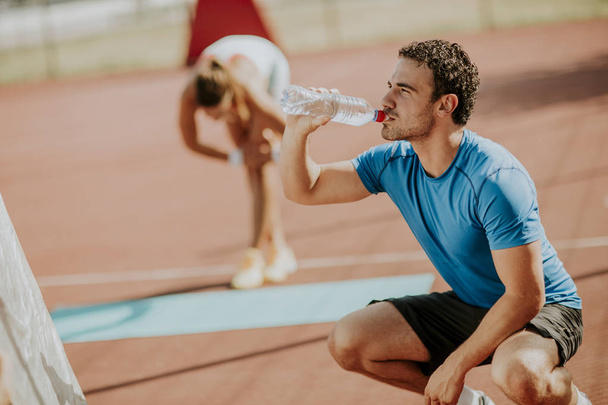 Sporty man drinking water while young woman doing exercise in the background outdoor - Zdjęcie, obraz