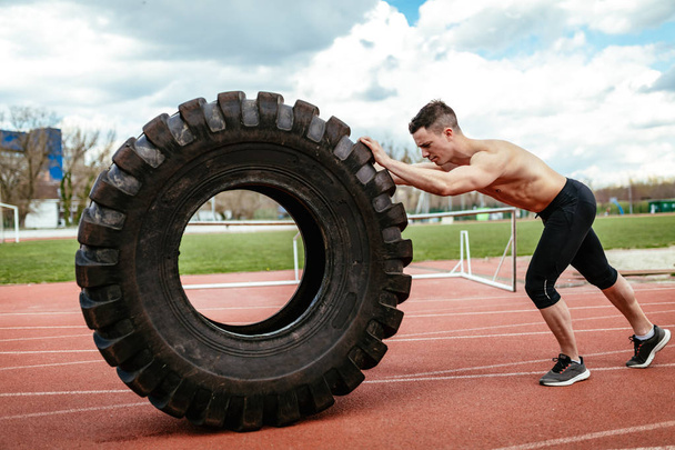 Молодой мускулистый человек поднимает огромные шины для тренировки мышц на стадионе
 - Фото, изображение