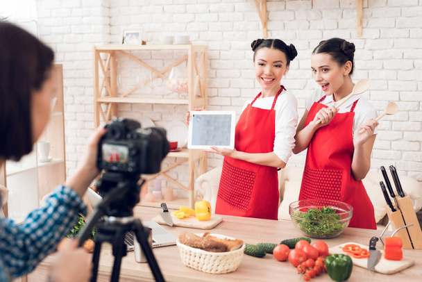 Two culinary bloggers in red aprons hold up tablet and spatulas to camera. - Foto, Imagem