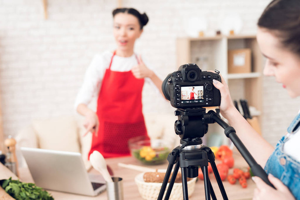 Two culinary bloggers in red aprons using laptop, one girl behind camera - Foto, Imagen