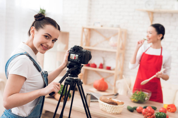 Two female culinary bloggers in red aprons tasting salad, one girl behind camera - 写真・画像