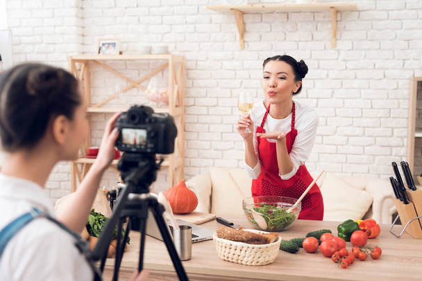 Two female culinary bloggers in red aprons tasting wine, one girl behind camera - 写真・画像