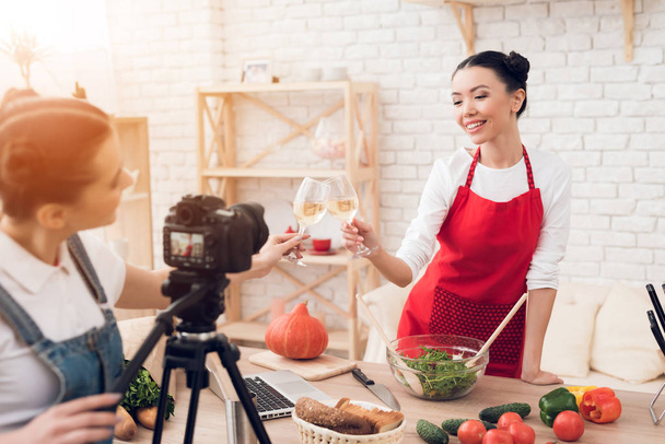 Two female culinary bloggers in red aprons tasting wine, one girl behind camera - Zdjęcie, obraz