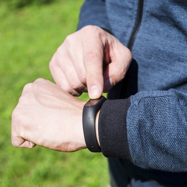 Fitnessarmband oder Smartwatch an der Hand eines Mannes. vor dem Hintergrund des grünen Rasens. ein erwachsener Kater mit blauem Band. Konzept eines gesunden Lebensstils - Foto, Bild