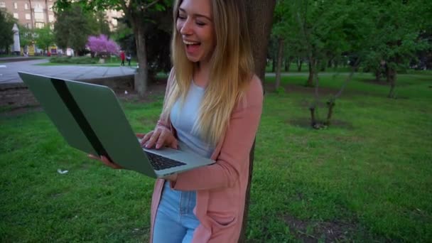 Jocund tyttö selailu kannettavan tietokoneen ja nojaten puuhun keväällä puistossa
. - Materiaali, video