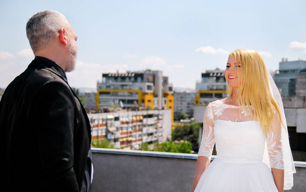 Wedding concept. Young happy handsome wedding couple stands on the roof of the building. Lovely bride is smiling and looking in the groom. In background are the buildings, the urban scene of the city. - Photo, image