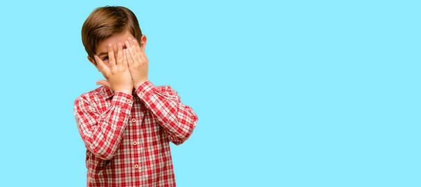 Handsome toddler child with green eyes smiling having shy look peeking through fingers, covering face with hands looking confusedly broadly over blue background - Photo, Image