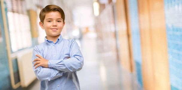 Όμορφος νήπιο παιδί με πράσινα μάτια με σταυρωμένα χέρια αυτοπεποίθηση και χαρούμενοι με ένα μεγάλο φυσικό χαμόγελο γελώντας στο διάδρομο του σχολείου - Φωτογραφία, εικόνα