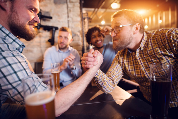 nuoret onnelliset parrakas miehet juovat olutta pubissa ja ottaa kädenvääntö haaste
  - Valokuva, kuva