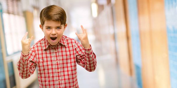 Criança bonita com olhos verdes fazendo símbolo de rocha com as mãos, gritando e celebrando no corredor da escola
 - Foto, Imagem