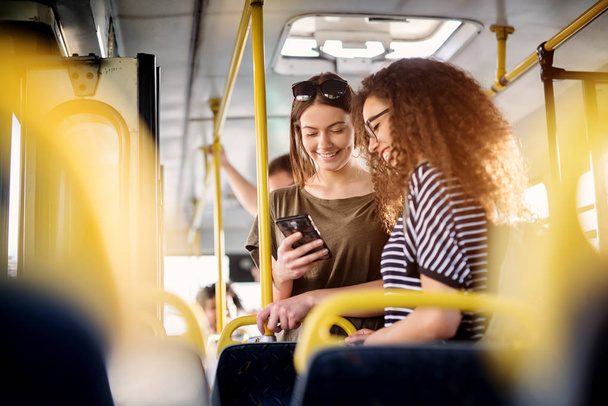 two cheerful pretty young women standing in a bus and looking at phone  - Zdjęcie, obraz