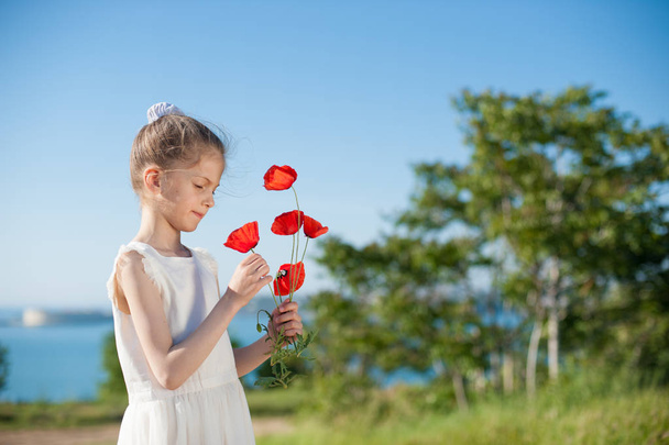 cute little girl in white dress holding bouquet of red poppy standing on sea bay background - Fotó, kép
