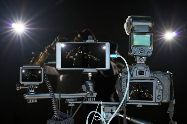 Professionelles Kameraset mit Smartphone und Action-Kamera auf Stativ über Musiker-Duo-Band, die Trompete mit Spotlicht und Linsenblitz auf der Bühne spielt. Live-Streaming für musikalisches Konzept - Foto, Bild