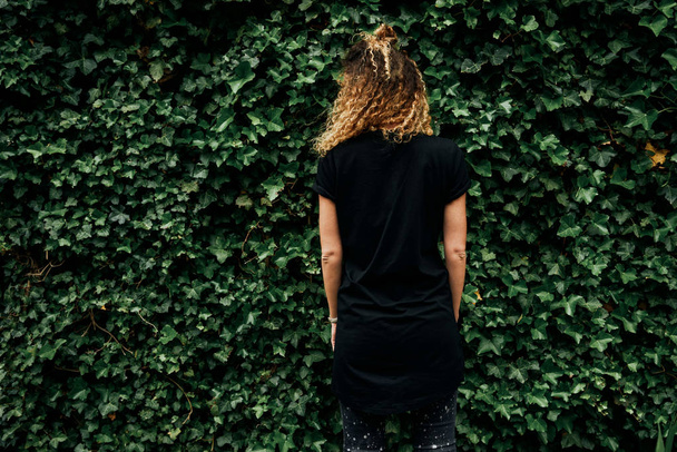 Young hipster woman wearing blank black t-shirt and black jeans standing on a plants background  - Fotó, kép