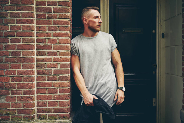 handsome hipster man wearing blank gray t-shirt standing against entrance door on a street  - Фото, зображення