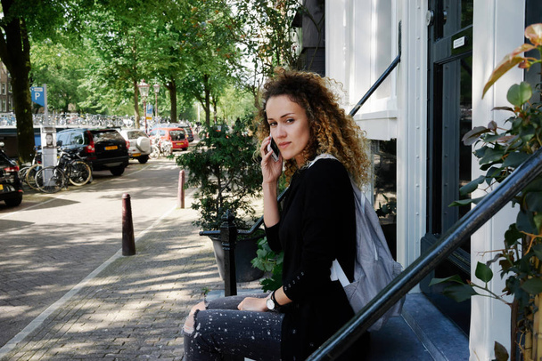 attractive young woman with curly hair talking on smartphone while sitting in a cozy city street - Foto, Bild