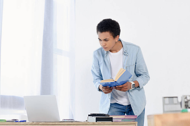 african american teenager holding book and looking at laptop at home - Fotoğraf, Görsel