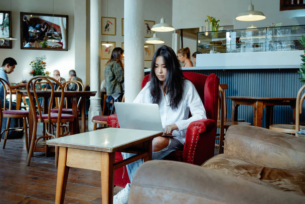 コーヒー ショップで距離ジョブのラップトップを使用してカジュアルなアジアの若い女性 - 写真・画像