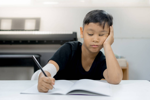 ein asiatischer Junge an den verwundeten Händen ist es leid, ein Notizbuch zu schreiben - Foto, Bild