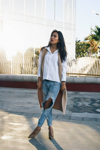 Young asian woman wearing white shirt, blue jeans and a long waistcoat is standing in a daylight on a street against fountain - Φωτογραφία, εικόνα