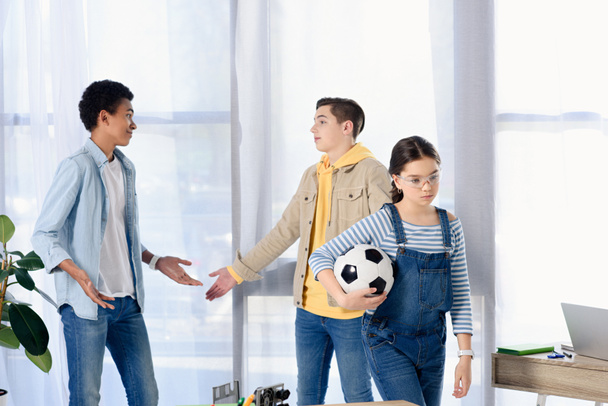 multicultural teenagers showing shrug gestures while friends walking away with football ball at home - Foto, Bild