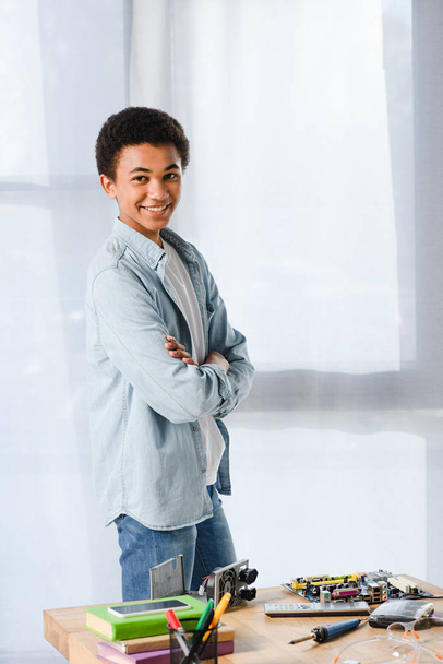 sonriente afroamericano adolescente de pie con los brazos cruzados cerca de la mesa con el equipo en casa
 - Foto, Imagen